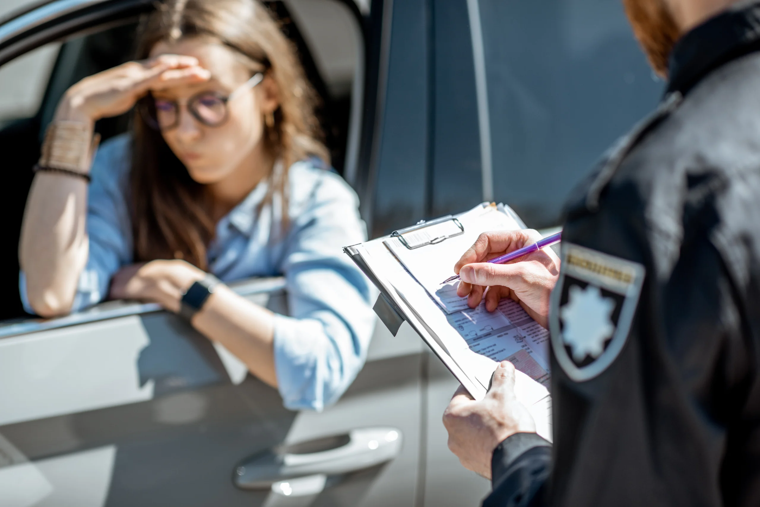 woman sitting in her car holding her head while a police officer writes a ticket